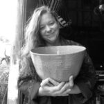 Black & white image of a white woman with long. hair smiling and holding a large ceramic bowl.